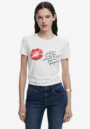 T-shirt en coton blanc avec une empreinte de rouge à lèvres rouge et un imprimé texte noir. Design ajusté avec des manches courtes, associé à un jean en denim bleu.