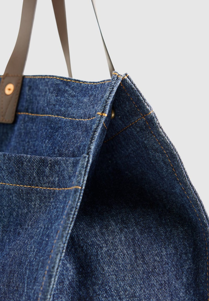 Jeanstasche mit braunen Lederriemen, sichtbaren Nähten und offenem Oberteil vor einem schlichten weißen Hintergrund.