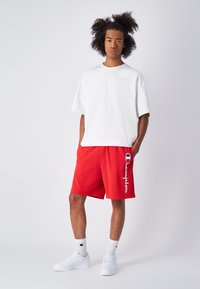 Camiseta blanca de manga corta, pantalones cortos deportivos rojos con el logo de "Champion" y zapatillas blancas. Los pantalones cortos tienen una textura suave y un corte relajado.