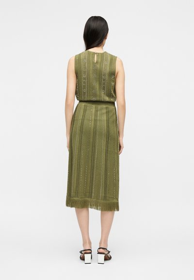 Γυναίκα με μακριά σκούρα μαλλιά, φοράει ένα ελαιί πράσινο αμάνικο φόρεμα με κάθετα σχέδια και φρατζόλες στο τελείωμα, στέκεται με την πλάτη στην κάμερα, μαύρα σανδάλια.