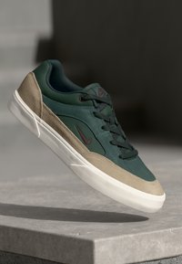 Green and tan sneaker with suede and leather materials, featuring a rubber sole, round laces, and a small maroon logo.