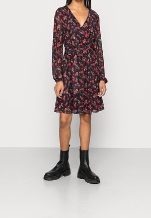 Mujer con vestido negro de manga larga con estampado floral rojo y botines negros, de pie frente a un fondo liso gris claro.