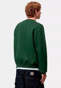 Sudadera verde oscuro con cuello redondo, hombros caídos y puños acanalados. Lleva puesta sobre una camisa blanca y combinada con jeans oscuros.
