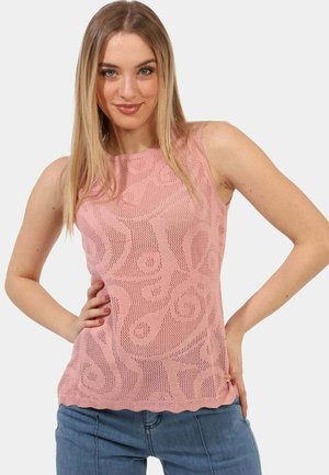 Giovane donna con lunghi capelli biondi, indossa un top rosa senza maniche a fantasia e jeans blu, posando con una mano sui fianchi.