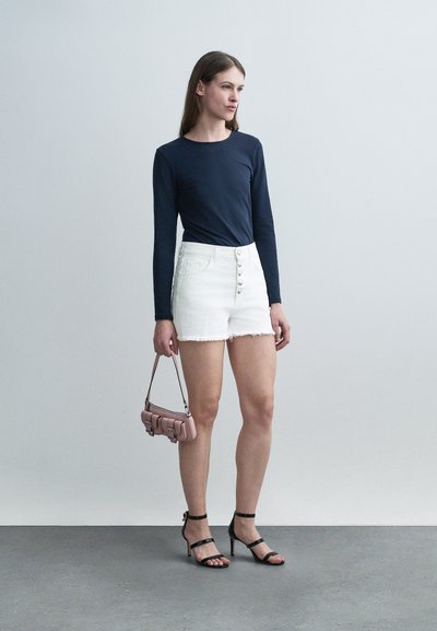 Γυναίκα που στέκεται με λευκό σορτς με κουμπιά μπροστά, μπλε μακρυμάνικο τοπ, μαύρα πέδιλα με τακούνι, κρατώντας μια μικρή ροζ τσάντα στο πλάι της.