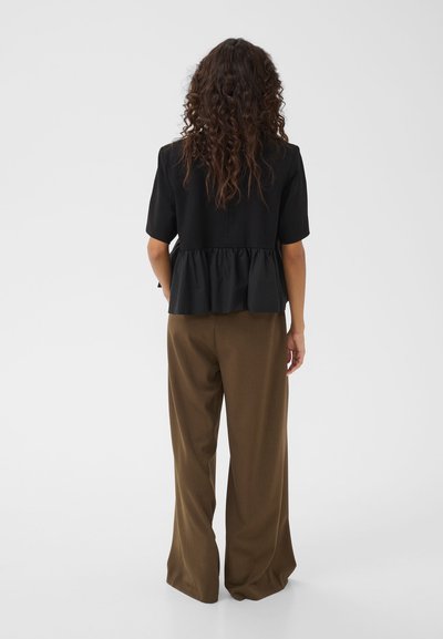 Femme aux longs cheveux bouclés, portant un haut noir à manches courtes et un pantalon marron ample, debout de dos sur un fond blanc uni.
