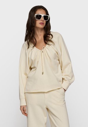 Kobieta w oversize’owych białych okularach przeciwsłonecznych, luźnej kremowej bluzce ze ściągaczem przy dekolcie i pasujących spodniach, pozująca na jednolitym tle.