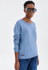 Jasnoniebieski sweter z miękkiej dzianiny, z okrągłym dekoltem, ściągaczami na rękawach oraz małą brązową skórzaną naszywką z logo na dolnym końcu.