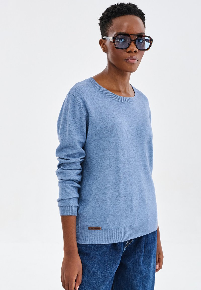 Jasnoniebieski sweter z miękkiej dzianiny, z okrągłym dekoltem, ściągaczami na rękawach oraz małą brązową skórzaną naszywką z logo na dolnym końcu.