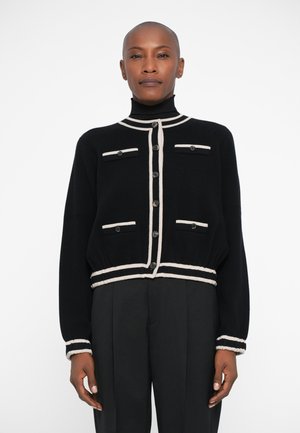 Schwarze Strickjacke mit beigen Akzenten, die einen hohen Kragen, eine Knopfleiste und zwei Brusttaschen mit Knöpfen auf jeder Seite hat. Weicher Stoff.
