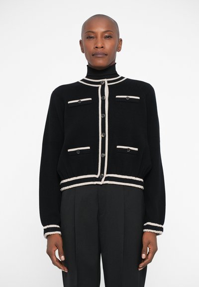 Sort cardigan med beige accenter, der har en høj krave, knappelukning foran og to brystlommer med knapper på hver side. Blød tekstur.