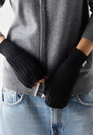 Black knitted fingerless gloves worn with grey zip-up sweater and light blue jeans, hands loosely clasped at waist.