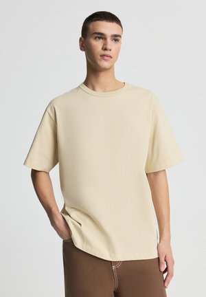 Maglietta beige a maniche corte in morbido cotone, con scollo rotondo e vestibilità rilassata, abbinata a pantaloni marroni dalla vestibilità comoda.