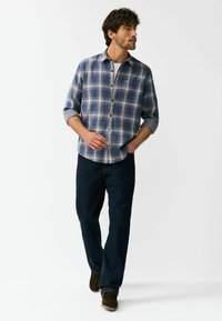 Kék kockás flanel ing barna gombokkal, feltűrt ujjal és egy mellzsebbel, sötét farmernadrággal és barna cipővel kombinálva.