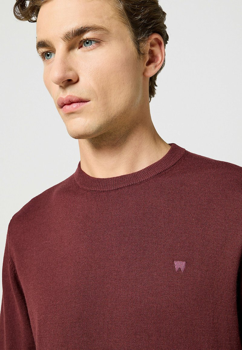 Maglione a maglia bordeaux con scollo rotondo, caratterizzato da un piccolo logo viola sul petto. La texture appare liscia e leggera.