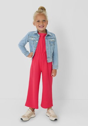 Jeune fille aux cheveux blonds attachés en chignon, portant une veste en jean bleu clair, une combinaison rouge vif et des baskets beige, souriant avec la main sur la hanche.