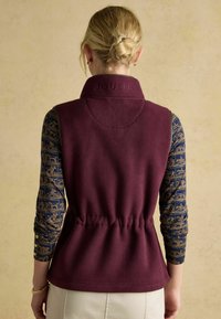 Burgunderfarbener Fleece-Weste mit hohem Kragen, elastischem Bund und geprägtem "JOULES"-Logo. Darunter ist ein gemustertes Langarmshirt.