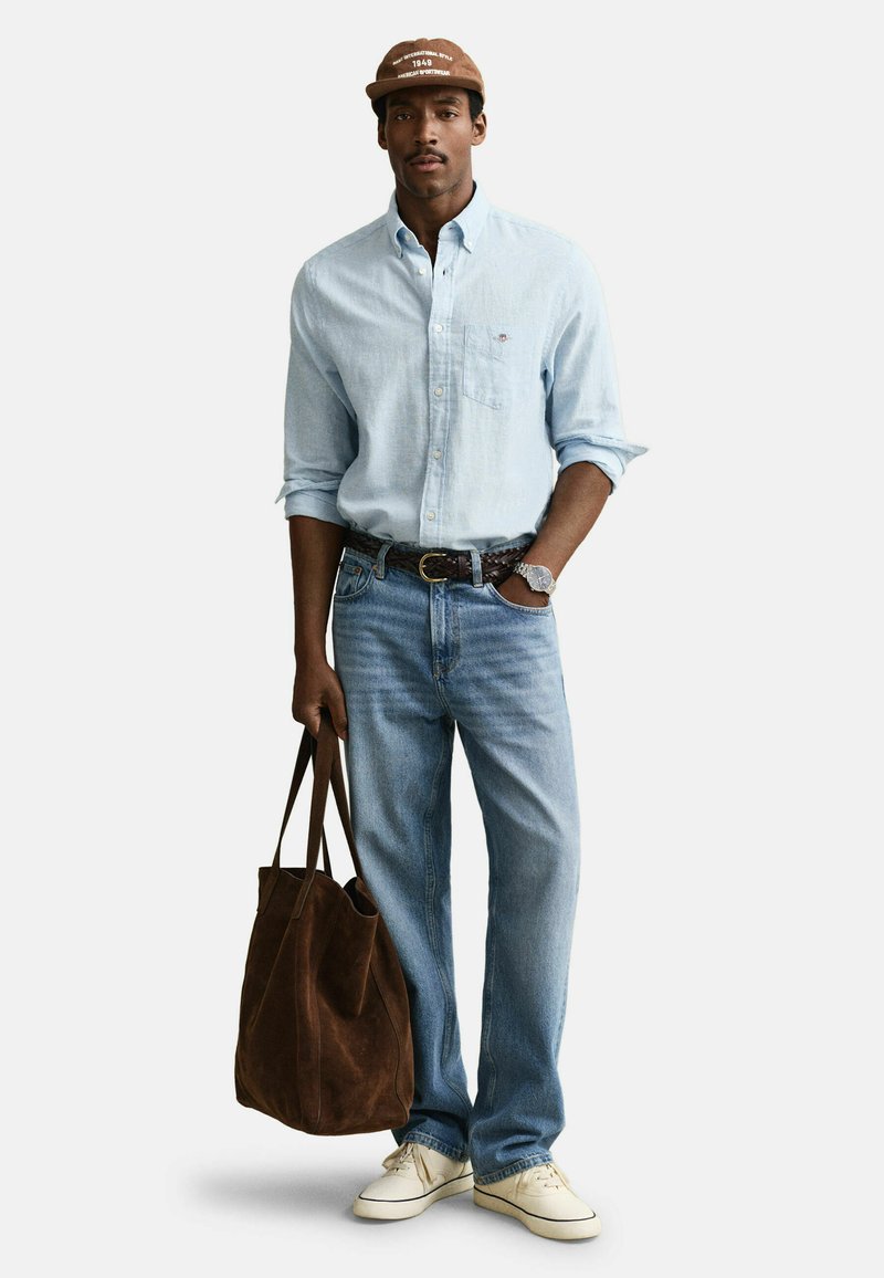 Mand iført lyseblå skjorte med knapper, blå jeans, hvide sneakers, brun kasket og ur, holder stor brun tote-taske ved siden.