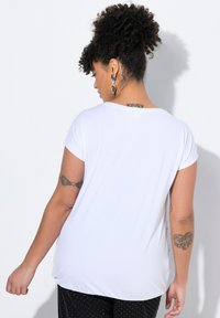 Camiseta blanca de manga corta con un corte holgado, escote redondo y textura suave, combinada con pantalones de patrón de puntos. La vista desde atrás muestra el detalle del tatuaje.