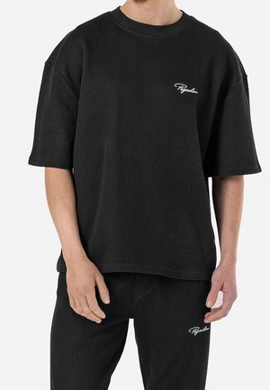 Mann trägt schwarzes, geripptes Kurzarm-T-Shirt und passende Hose mit kleinen weißen "Pegador"-Textlogos auf Brust und Oberschenkel.