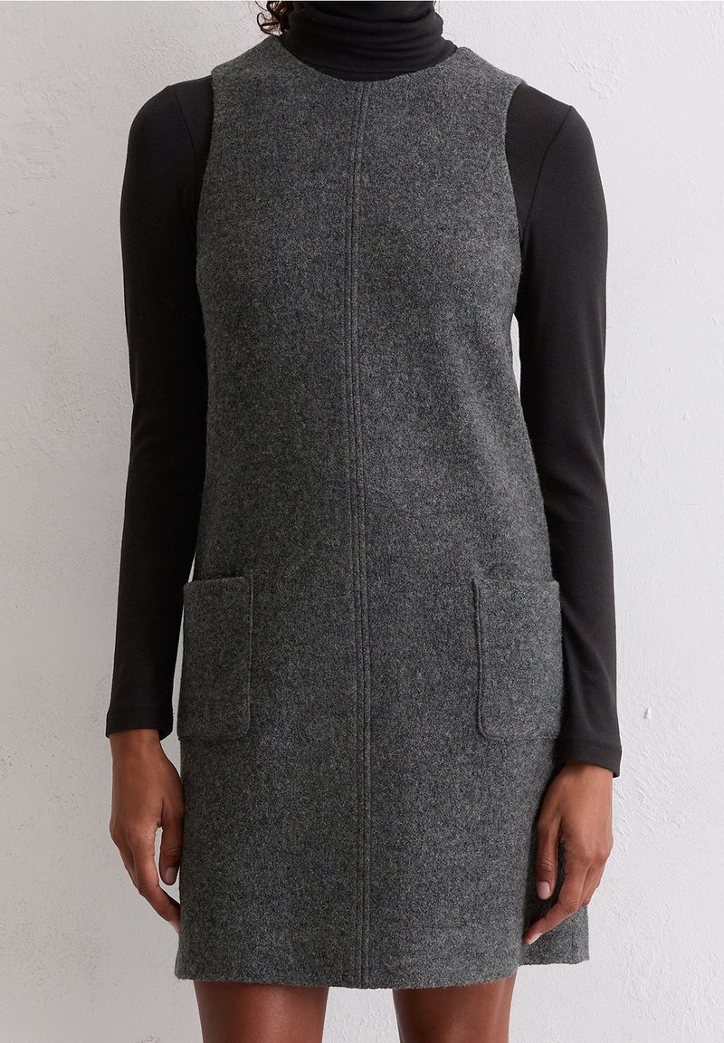 Femme portant une robe sans manches en laine grise avec des poches avant sur un haut noir à manches longues et col roulé.