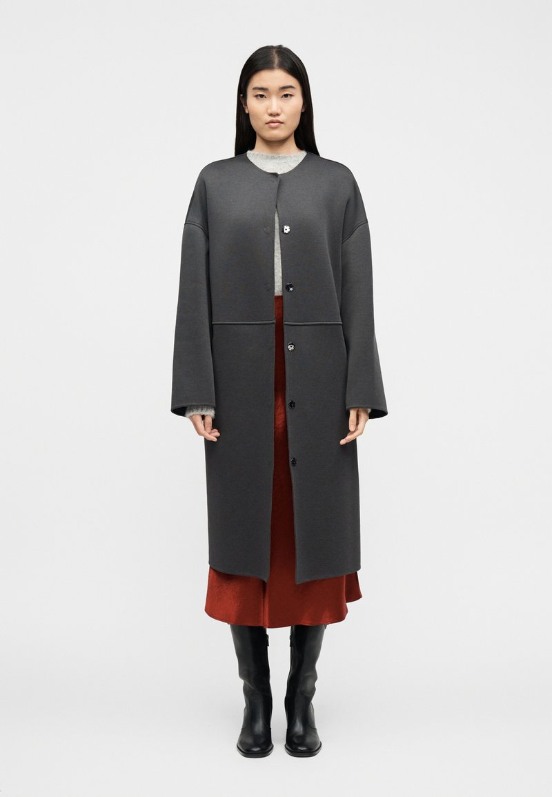 Γυναίκα που φοράει μακρύ σκουρόχρωμο γκρι παλτό με κουμπιά, ανοιχτό γκρι πουλόβερ, φούστα σε σκουριά απόχρωση και μαύρες μπότες έως το γόνατο, στέκεται μπροστά σε λευκό φόντο.