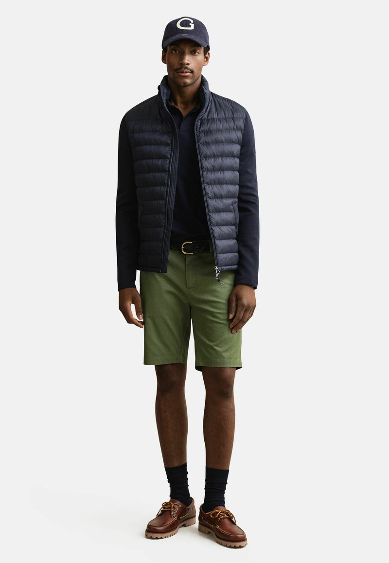 Mann trägt marineblaue Mütze, schwarze Steppjacke über schwarzem Langarmshirt, olivgrüne Shorts, schwarze Socken und braune Lederschuhe.
