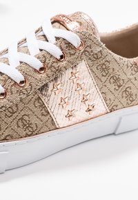 Béžová látková teniska s bílými tkaničkami, kovovými růžově zlatými akcenty, hvězdnými detaily a bílou gumovou podrážkou. Viditelné strukturované povrchy a vzory.