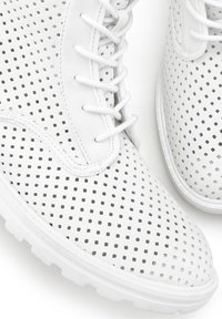 Bílé perforované tenisky s texturovaným povrchem, vyznačující se plochými bílými tkaničkami a silnou gumovou podešví. Design zahrnuje prodyšný vzor.