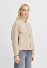 Giacca bomber beige con cerniera frontale, tasche laterali e colletto a costine. Realizzata in tessuto morbido, abbinata a jeans a gamba larga grigio chiaro.