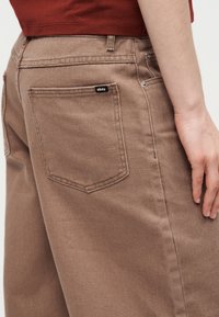 Κοντινό πλάνο ενός ατόμου που φοράει ανοιχτό καφέ τζιν παντελόνι με ορατή πίσω τσέπη που φέρει την επιγραφή «obey» και κόκκινο πουκάμισο μέσα από το παντελόνι.