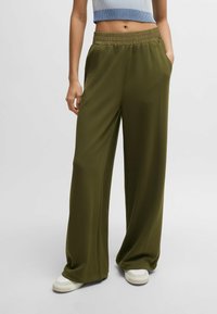 Oliven grüne weit geschnittene Hose mit elastischem Bund, aus weichem Stoff und seitlichen Taschen. Kombiniert mit weißen Sneakers, für einen bequemen Look gestylt.