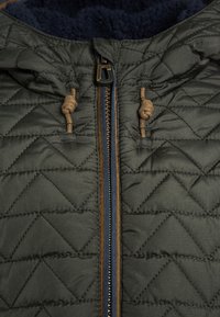 Kviltet grøn jakke med marineblå fleeceforing, frontlynlås med brune detaljer og snore i hætten. Struktureret overflade.