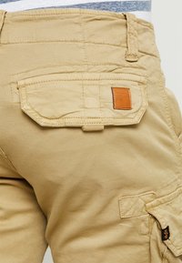 Shorts cargo beiges avec poches à rabat et fermetures à bouton, ainsi qu'une petite étiquette de marque en cuir marron sur la poche arrière.