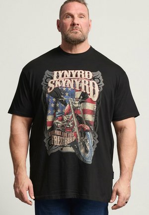 Mann trägt schwarzes T-Shirt mit Lynyrd Skynyrd Schriftzug, rotem Motorrad und amerikanischer Flagge im Hintergrund.