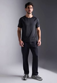 Črna športna majica s kratkimi raglan rokavi, lahka tkanina; v kombinaciji s črnimi hlačami. Model stoji na nevtralni podlagi.