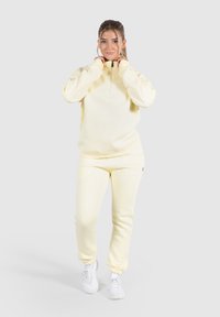 Ljust gul zip-up sweatshirt med hög krage, parat med matchande avsmalnande sweatpants. Modellen står med händerna nära kragen, iförd vita skor.