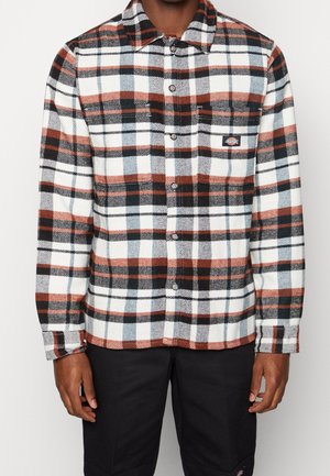 Homme portant une chemise à carreaux boutonnée à manches longues aux couleurs noir, blanc et rouille avec une poche poitrine.