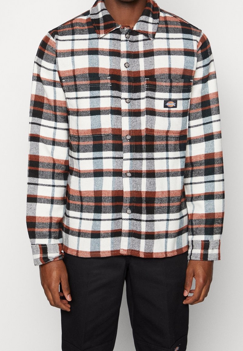 Homme portant une chemise à carreaux boutonnée à manches longues aux couleurs noir, blanc et rouille avec une poche poitrine.