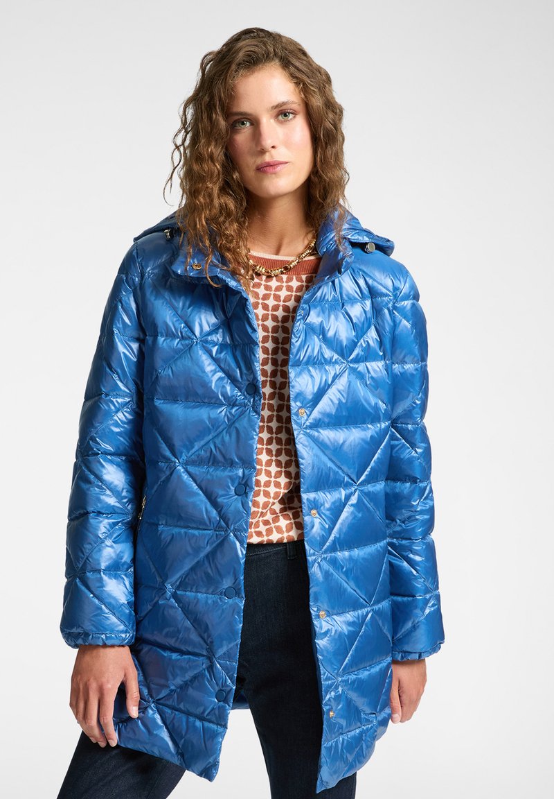 Blauwe pufferjas met een glanzend, gestikt uiterlijk en een kap. Heeft een knoopsluiting en gouden accenten. Gekleed met een gebreide trui met patroon.