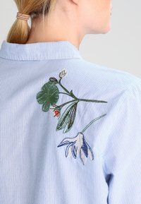 Camicia a righe blu chiaro con colletto, con dettagli di ricami floreali in verde, bianco e blu sulla schiena.
