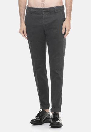 Uomo in pantaloni grigi scuri con risvolto e scarpe eleganti nere lucide, in piedi davanti a uno sfondo bianco uniforme.
