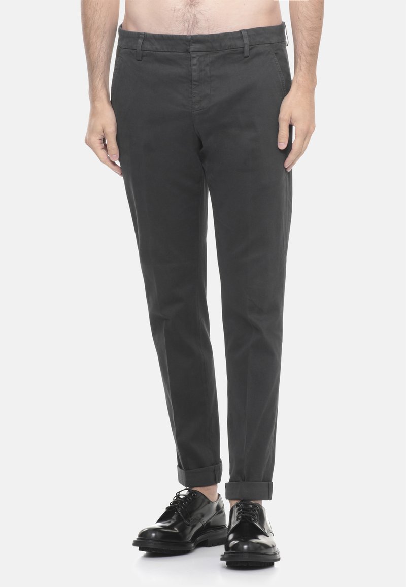 Man in een donkergrijs opgerolde broek en glanzende zwarte veterschoenen, staande tegen een effen witte achtergrond.