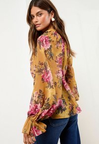 Blumenbluse in Senfgelb mit rosa und braunen Blumen. Verfügt über Puffärmel mit elastischen Bündchen und einem weichen, leichten Stoff.