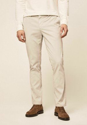 Mężczyzna noszący beżowe wąskie spodnie oraz brązowe zamszowe wiązane buty, stojący na tle jasnego, jednolitego tła.