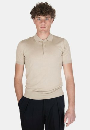 Giovane uomo con capelli ricci che indossa una polo beige a maniche corte e pantaloni neri, in piedi con le mani in tasca su sfondo bianco.