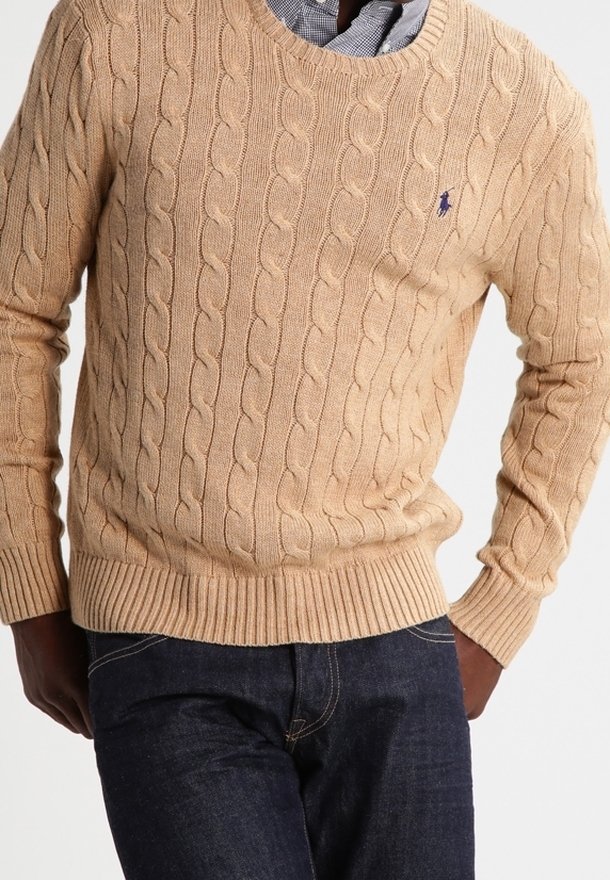 Homme portant un pull beige à torsades par-dessus une chemise à carreaux bleue, associé à un jean bleu foncé, debout les mains dans les poches.