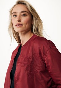 Veste bomber bordeaux avec un design floral brodé sur la poitrine gauche, dotée d'une fermeture éclair à l'avant et d'un col en côtes. Texture lisse, coupe décontractée.