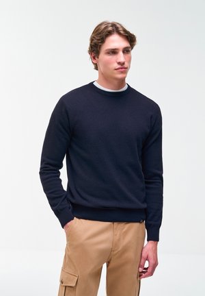 Jonge man met een marineblauwe trui en beige cargobroek, staand met één hand in zijn zak tegen een effen witte achtergrond.