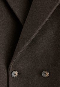Tessuto in misto lana marrone con una texture liscia, presenta un design doppiopetto con due bottoni. I rever sartoriali mettono in risalto il capo.